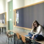 A woman sits on a desk in a university hallway, studying on a laptop.