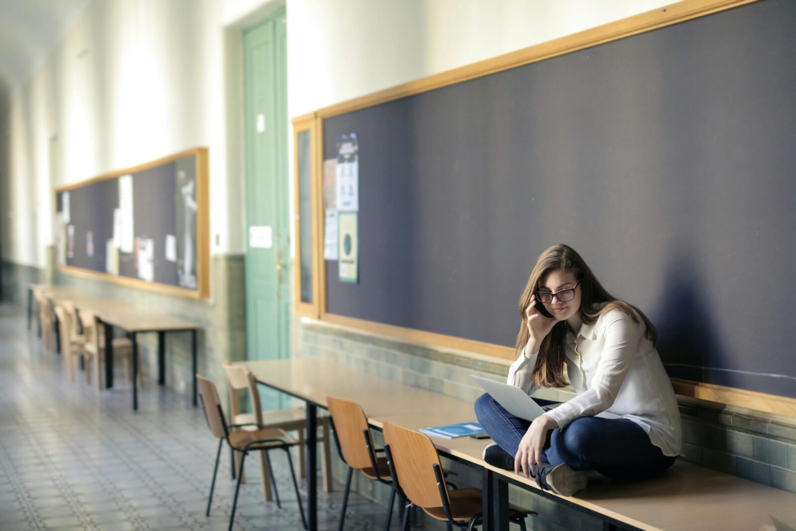 A woman sits on a desk in a university hallway, studying on a laptop.